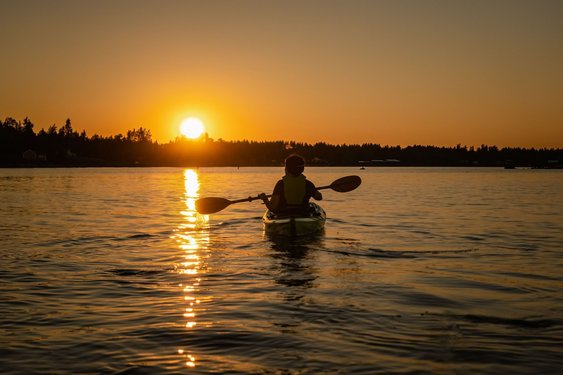 Auf einer Wasserlandschaft im Vordergrund paddelt jemand in einem Kanu, während im Hintergrund gelb-orange die Sonne hinter Bäumen untergeht.