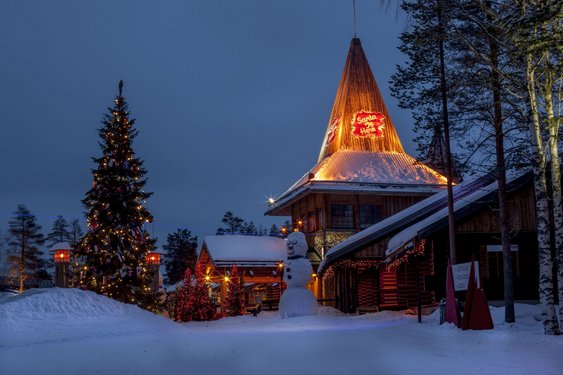 Ein mit Lichterketten geschmücktes Haus steht flankiert von einem Weihnachtsbaum in einer Schneelandschaft.