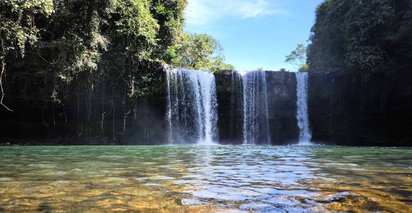 Über eine klare Wasserfläche schaut man auf einen links und rechts von üppig-grüner Vegetation umgebenen Wasserfall.