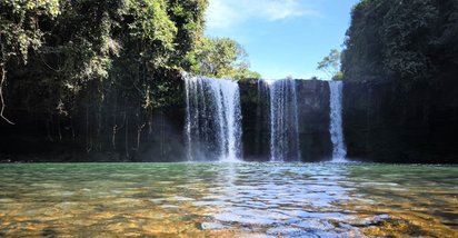 Tad-Champee-Wasserfall am Bolaven-Plateau Über eine klare Wasserfläche schaut man auf einen links und rechts von üppig-grüner Vegetation umgebenen Wasserfall.