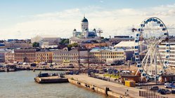 Vom Wasser im Hafen aus blickt man auf die Skyline Helsinkis. Zentral hinten im bild befindet sich der weiße Dom, rechts im Vordergrund steht ein Riesenrad.