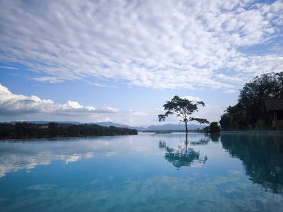 Im Vordergrund sieht man eine weite glatte Wasserfläche, die im Bildmittelgrund von Bäumen und Bergen begrenzt wird. Die obere Bildhälfte wird von blauem Himmel mit dichten, weißen Wolken eingenommen.