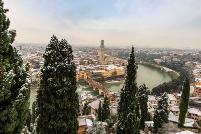 Über Zypressenwipfel im Vordergrund schaut man auf das von der Etsch durchzogene Verona. Auf den Bäumen und Dächern liegt eine leichte Schneedecke.