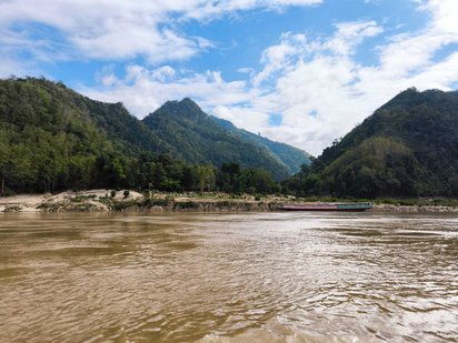 Das untere Drittel des Bildes wird von einer braunen Wasserfläche eingenommen. Am dahinter befindlichen Ufer erheben sich bewaldete Berge.