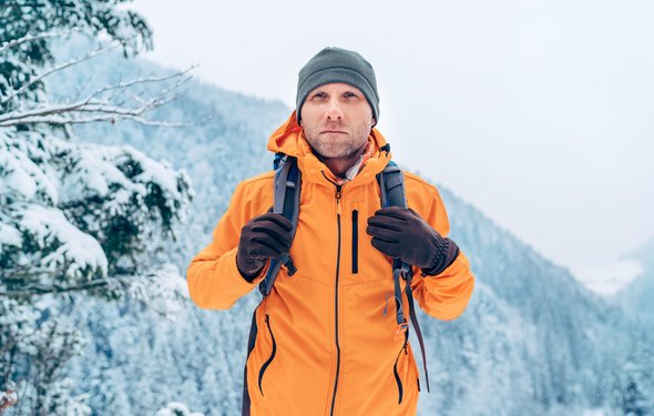 Ein Wanderer in einer orangenen Outdoor-Jacke läuft durch eine verschneite Waldlandschaft, er steht vorne im Bild und schaut den Betrachter an.
