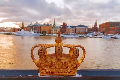 Im Vordergrund ziert eine große goldene Krone die Bildmitte. Dahinter erstreckt sich eine Wasserfläche, über die man auf am Kai liegende Boote und dahinter historische Gebäude blickt. An der Krone und auf den Dächern der Häuser befindet sich Schnee.