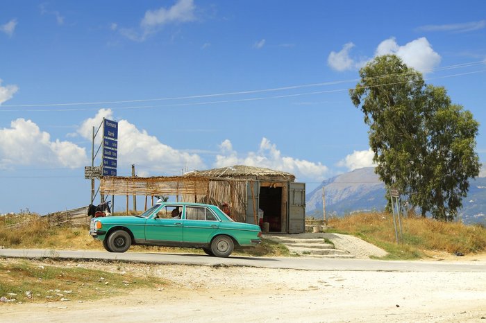 Blick in eine Landschaft in Albanien, im Vordergrund steht eine Hütte mit einem blauben Oldtimer davor.