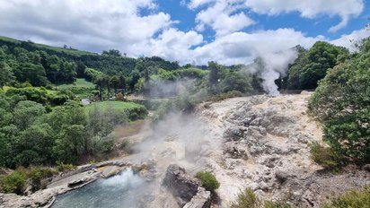 Im Bildvordergrund sieht man, wie aus zwischen Felsen gelegenen Wasserflächen Dampf aufsteigt. Rechts im Bild und hinter der Felsfläche erheben sich Bäume, darüber erstreckt sich ein von großen weiß-grauen Wolken bedeckter Himmel.