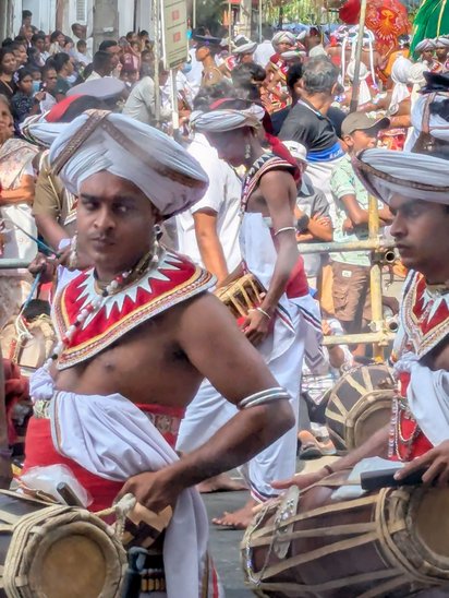 Sri-lankische Trommler in traditionellen Trachten ziehen über eine Straße und werden von einer großen Menschenmenge betrachtet.