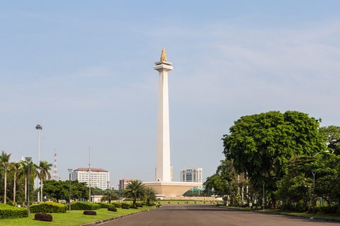 Das Nationaldenkmal Monas in Jakarta, Indonesien, steht inmitten einer großen Parkanlage. Die hohe, weiße Säule wird von einer goldenen Flamme gekrönt. Im Vordergrund führt eine breite Straße und grüne Bäume zum Denkmal.