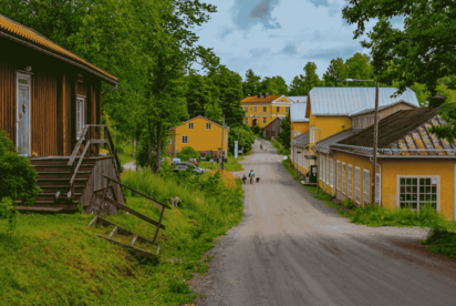 An einer nicht asphaltierten Straße liegen historische Gebäude: angeschnitten im Vordergrund links ein rotes Holzhaus, dahinter im Bildmittegrund mehrere gelbe Steinhäuser.
