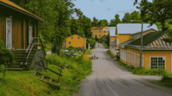 An einer nicht asphaltierten Straße liegen historische Gebäude: angeschnitten im Vordergrund links ein rotes Holzhaus, dahinter im Bildmittegrund mehrere gelbe Steinhäuser.