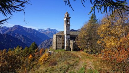 Ein Gras bewachsener Weg führt auf eine alte Kirche in der Bildmitte zu. Das Bild wird von herbstlich gefärbten Bäumen gerahmt. Hinter der Kirche erstreckt sich eine Berglandschaft.