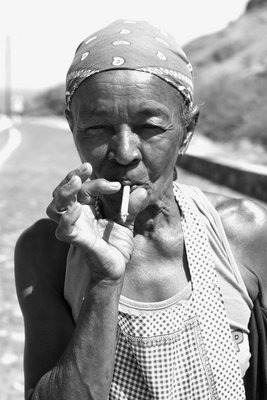 Schwarz-Weiß-Porträt einer alten rauchenden Kapverdierin. Sie trägt ein helles Tanktop, darüber eine Schürze sowie ein Kopftuch, so dass ihre Haare nicht zu sehen sind.