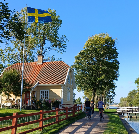 Im Garten eines beigen Holzhauses weht eine Schweden-Fahne, rechts fahren zwei Radfahrer:innen vom Betrachter weg, daneben ein Kanal.