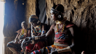 Im inneren einer Lehmhütte sitzen drei Afrikaner in traditioneller Kleidung auf einer Bank vor der Hüttenwand.