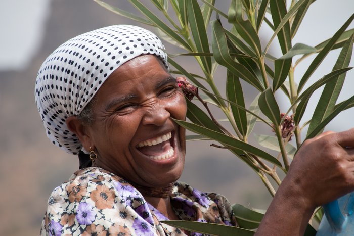 Lachende Frau mit weiß-schwarz gepunktetem Kopftuch hält einen Oleander-Zweig auf den Kapverden. 