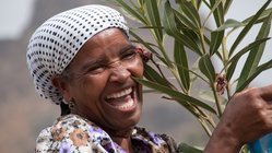 Lachende Frau mit weiß-schwarz gepunktetem Kopftuch hält einen Oleander-Zweig auf den Kapverden. 