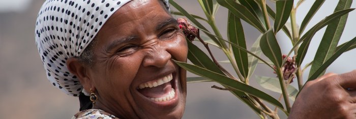 Lachende Frau mit weiß-schwarz gepunktetem Kopftuch hält einen Oleander-Zweig auf den Kapverden. 