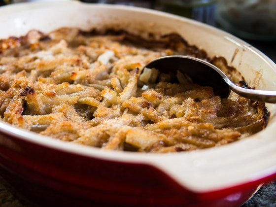 Janssons frestelse, schwedischer Kartoffelauflauf In einer roten Auflauffprm befindet sich ein knusprig überbackenes Gericht, in dem ein Löffel steckt.