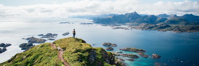 Ein Wanderer steht auf einem Berggipfel auf den Lofoten in Norwegen und blickt über eine beeindruckende Landschaft aus Bergen, Fjorden und Inseln.