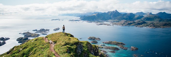 Ein Wanderer steht auf einem Berggipfel auf den Lofoten in Norwegen und blickt über eine beeindruckende Landschaft aus Bergen, Fjorden und Inseln.