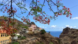 Oben links ragt ein Zweig mit roten Blüten ins Bild, links unten erheben sich bunte Häuser an einer Bergwand, rechths im Hintergrund sieht man durch eine Bergöffnung auf das Meer.