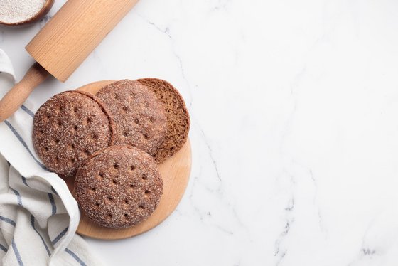Finnlands Nationalgericht, das Roggenbrot Ruisleipä Auf einer weißen Oberfläche steht links ein Holzbrett, auf dem sich vier der kleinen finnischen Roggenbrotfladen Ruisleipä befinden. Links neben dem Holzbrett sieht man im Anschnitt ein Nudelholz, ein blau-gestreiftes Küchentuch sowie eine Schale mit Mehl.