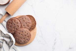 Finnlands Nationalgericht, das Roggenbrot Ruisleipä Auf einer weißen Oberfläche steht links ein Holzbrett, auf dem sich vier der kleinen finnischen Roggenbrotfladen Ruisleipä befinden. Links neben dem Holzbrett sieht man im Anschnitt ein Nudelholz, ein blau-gestreiftes Küchentuch sowie eine Schale mit Mehl.