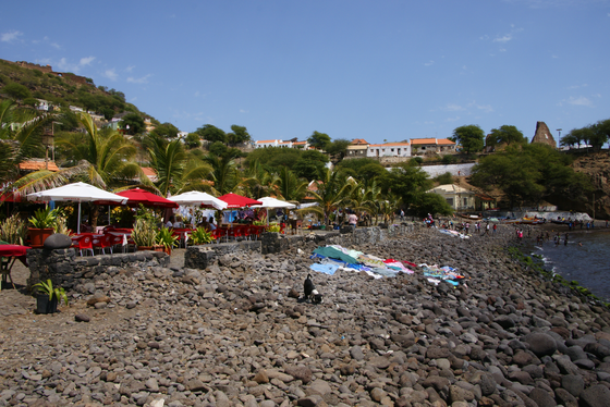 Auf dem Steinstrand trocknet Wäsche in der Sonne,entlang des Strandes stehen Stühle und Tische unter weißen und roten Sonnenschirmen. unter 