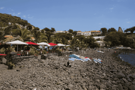 Auf dem Steinstrand trocknet Wäsche in der Sonne,entlang des Strandes stehen Stühle und Tische unter weißen und roten Sonnenschirmen. unter 