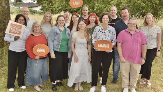 Das Team von REISEN MIT SINNEN steht freundlich lächelnd auf einer Wiese vor einem Waldstück in Dortmund.