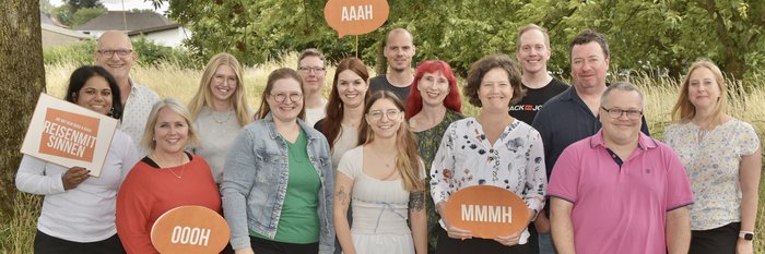 Das Team von REISEN MIT SINNEN steht freundlich lächelnd auf einer Wiese vor einem Waldstück in Dortmund.