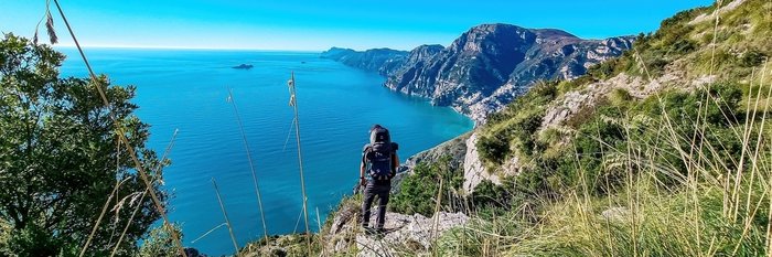 Rückenansicht eines Wanderers, der mit seinem Wanderrucksack auf einem Felsen steht und den Blick auf das blau schimmernde Meer gerichtet hat.