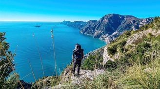 Rückenansicht eines Wanderers, der mit seinem Wanderrucksack auf einem Felsen steht und den Blick auf das blau schimmernde Meer gerichtet hat.