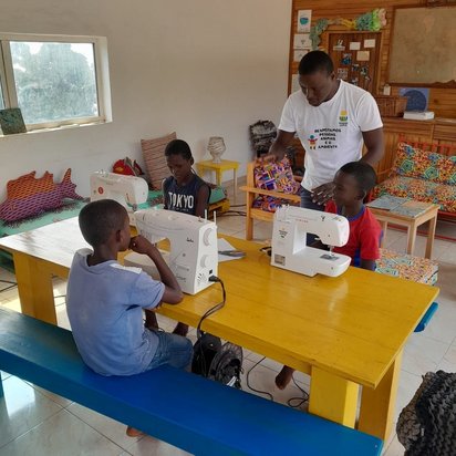 An einem gelben Tisch sitzen drei afrikanische Jungen vor weißen Nähmaschinen. Ein afrikanischer Mann steht neben ihnen und redet mit ihnen.