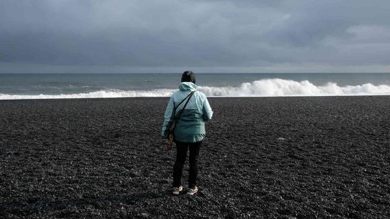 Spaziergang am schwarzen Strand von Reynisfjara Eine Person in türkisfarbener Jacke steht mit dem Rücken zur Kamera auf dem schwarzen Lavasand des Strands von Reynisfjara. Im Hintergrund brechen kräftige Wellen unter grauem Himmel.