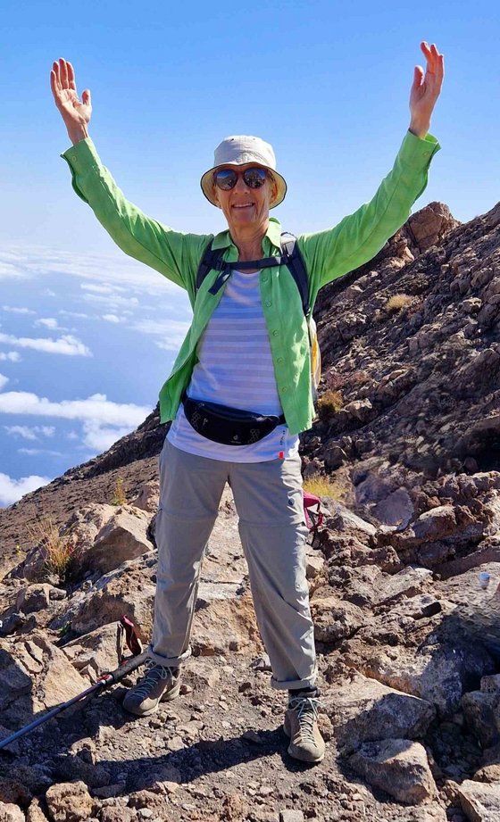 Eine Frau in Outdoor-Kleidung mit einer grasgrünen Jacke, Sonnenbrille und Mütze reckt freudig die Arme in die höhe. Sie steht auf einem felsigen Berggipfel, hinter ihr kann man den Abgrund erkennen. Der Himmel ist blau.