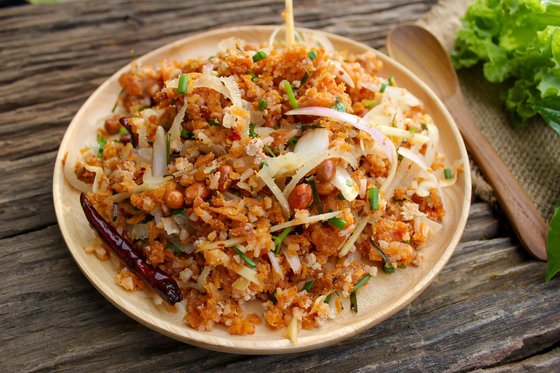 Ein Teller mit Naem Khao Tod, einem traditionellen laotischen Gericht aus knusprigem Reis, serviert mit frischen Kräutern, Limettenscheiben und Chilis, arrangiert auf einem Holztisch.