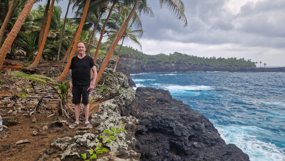 Dennis an der Küste von São Tomé Ein junger Mann steht unter Palmen an einer Felsküste, rechts liegt das blaue Meer.