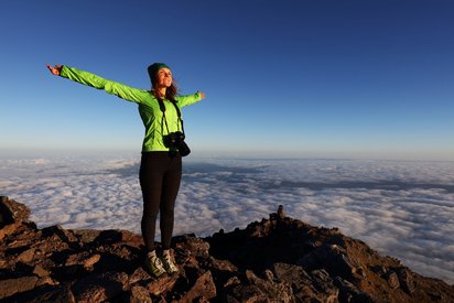 Azoren im Fokus Eine Frau in einer grünen Jacke und einer Kamera um den Hals steht mit ausgebreiteten Armen über dem Wolkenmeer auf einem Azoren-Gipfel.