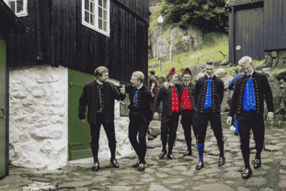 Eine Gruppe von Männern in färöischer Volkstracht läuft eine mit Steinplatten belegte Straße, an deren Rand Häuser stehen, hinunter.