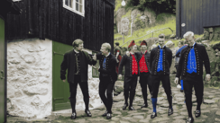 Eine Gruppe von Männern in färöischer Volkstracht läuft eine mit Steinplatten belegte Straße, an deren Rand Häuser stehen, hinunter.