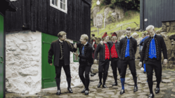 Eine Gruppe von Männern in färöischer Volkstracht läuft eine mit Steinplatten belegte Straße, an deren Rand Häuser stehen, hinunter.