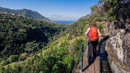 Frau in den Bergen auf einem ausgebauten Wanderpfad an einer Bergabhang neben einer Wasserstraße