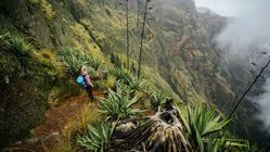 Aus der Vogelperspektive schaut man einen grün-bewachsenen Steilhang hinunter in einen Krater. Etwas links der Mitte steht eine blonde Frau mit einem blauen Wanderrucksack im Hang.