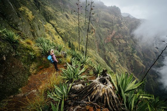 Wandern im Cova-Krater Aus der Vogelperspektive schaut man einen grün-bewachsenen Steilhang hinunter in einen Krater. Etwas links der Mitte steht eine blonde Frau mit einem blauen Wanderrucksack im Hang.