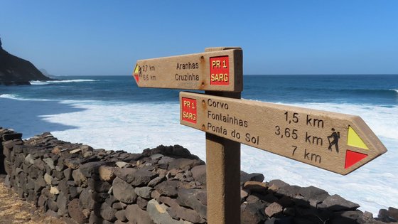 Santo Antão, das Wander-Eldorado der Kapverden Vor einer Mauer aus groben Natursteinen, hinter der sich das Meer befindet, steht ein Wanderwegweiser, der in zwei Richtungen die Distanzen zu Zielen nach links und nach rechts den Küstenwanderweg auf Santo Antao, der Wanderinsel der Kapverden, entlang angibt.