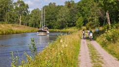 Rechts entlang eines Kanals verläuft ein gekiester Radweg. Im Hintergrund sind Radfahrer unterwegs, auf dem Kanal fährt ein Boot.