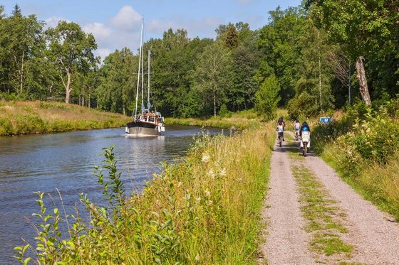 Rechts entlang eines Kanals verläuft ein gekiester Radweg. Im Hintergrund sind Radfahrer unterwegs, auf dem Kanal fährt ein Boot.